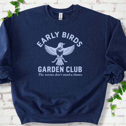 Navy crewneck sweatshirt laid flat on white brick with greenery. Pale blue bird holding worms between "Early Birds" and "Garden Club" chest lettering.