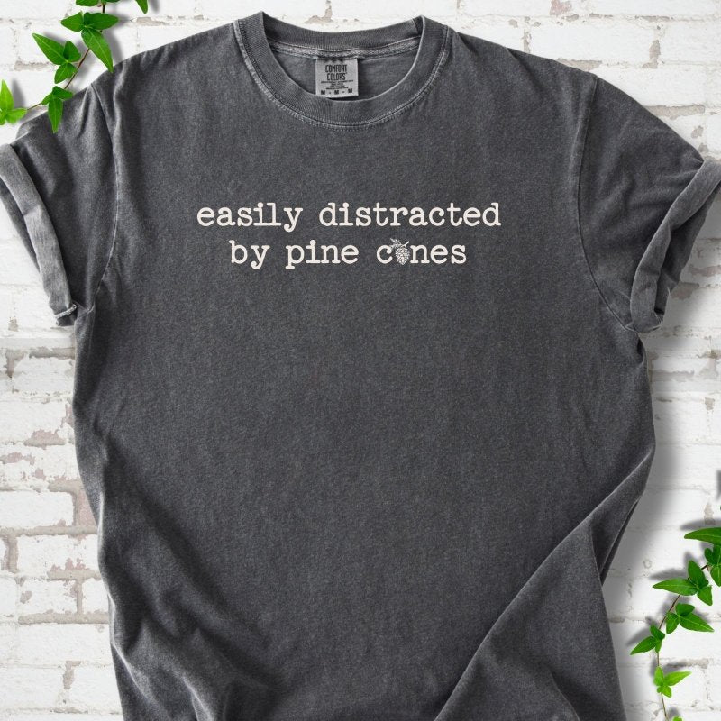 Pepper Comfort Colors® T-shirt shown flat with "Easily Distracted by Pine Cones" text and a small pine cone graphic