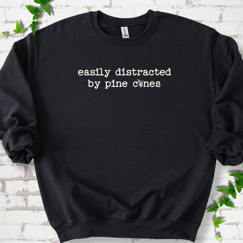 Black classic sweatshirt shown flat with "Easily Distracted by Pine Cones" text and a small pine cone graphic