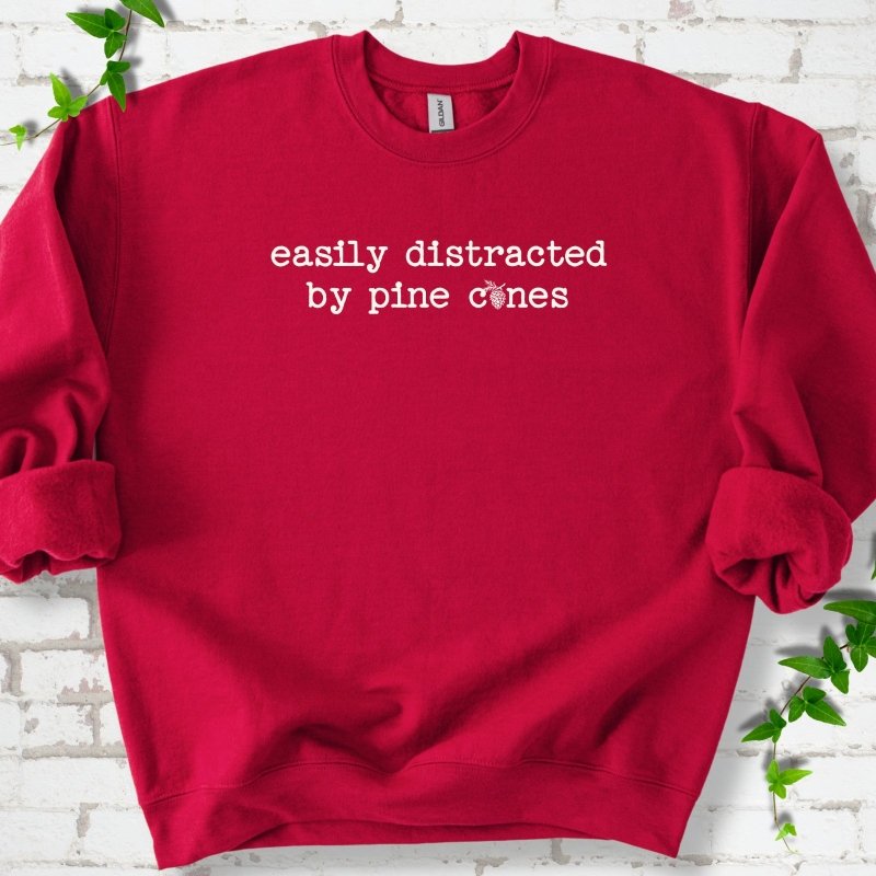 Cardinal Red classic sweatshirt shown flat with "Easily Distracted by Pine Cones" text and a small pine cone graphic