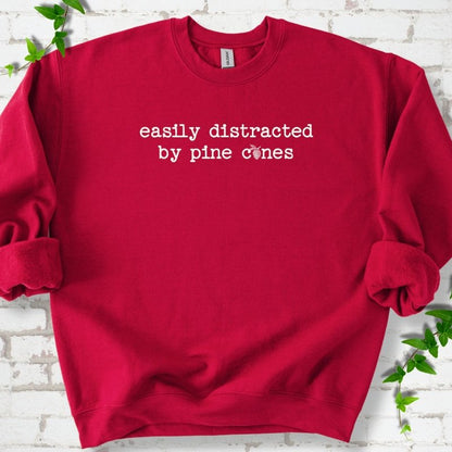Cardinal Red classic sweatshirt shown flat with "Easily Distracted by Pine Cones" text and a small pine cone graphic