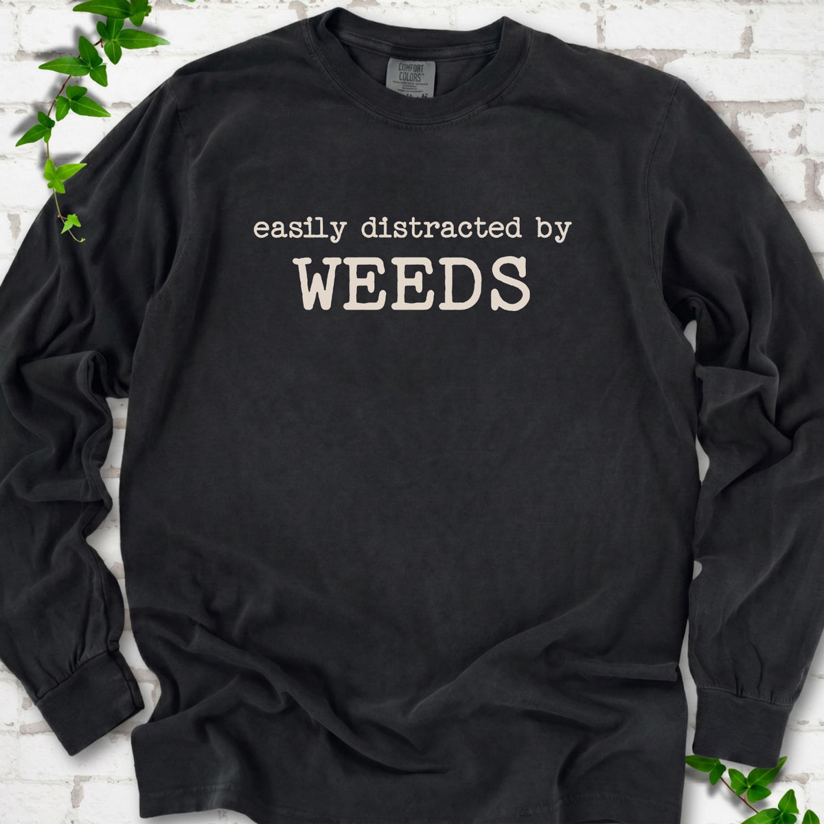 Black Comfort Colors® long sleeve T-shirt shown flat with cream "Easily Distracted by Weeds" gardening slogan