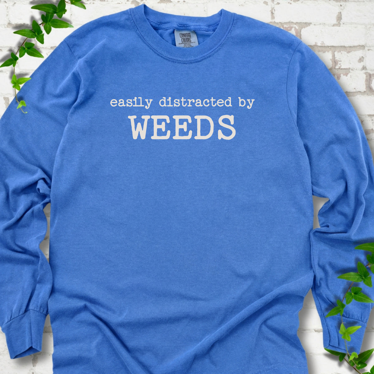 Flo Blue Comfort Colors® long sleeve T-shirt shown flat with cream "Easily Distracted by Weeds" gardening slogan
