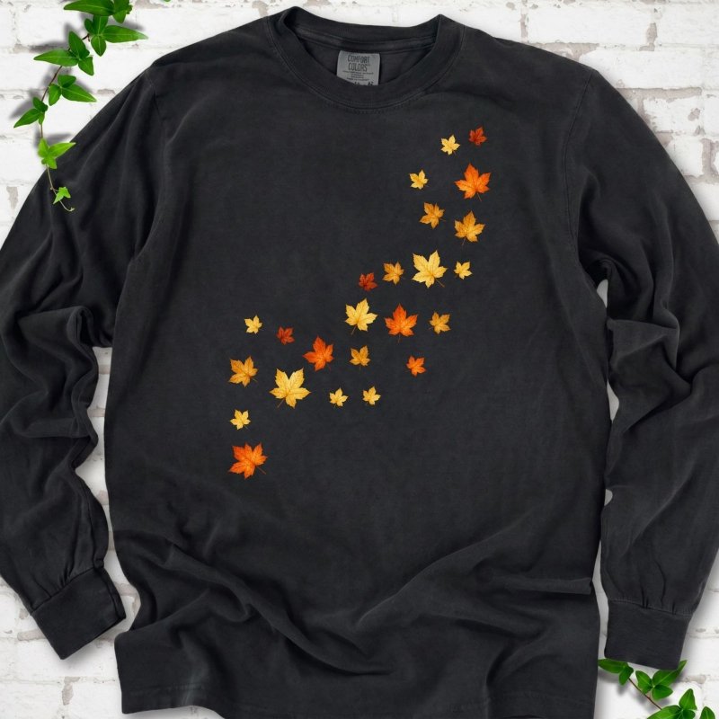 Black long sleeve t-shirt with a diagonal drift of small orange, gold and red maple leaves falling across the front, flat lay on white brick with ivy