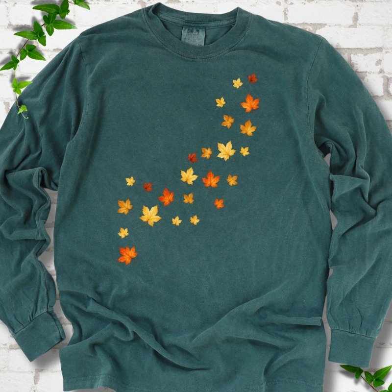 Blue Spruce deep teal long sleeve t-shirt with a diagonal drift of small orange, gold and red maple leaves falling across the front, flat lay on white brick with ivy