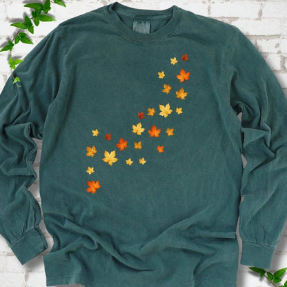 Blue Spruce deep teal long sleeve t-shirt with a diagonal drift of small orange, gold and red maple leaves falling across the front, flat lay on white brick with ivy