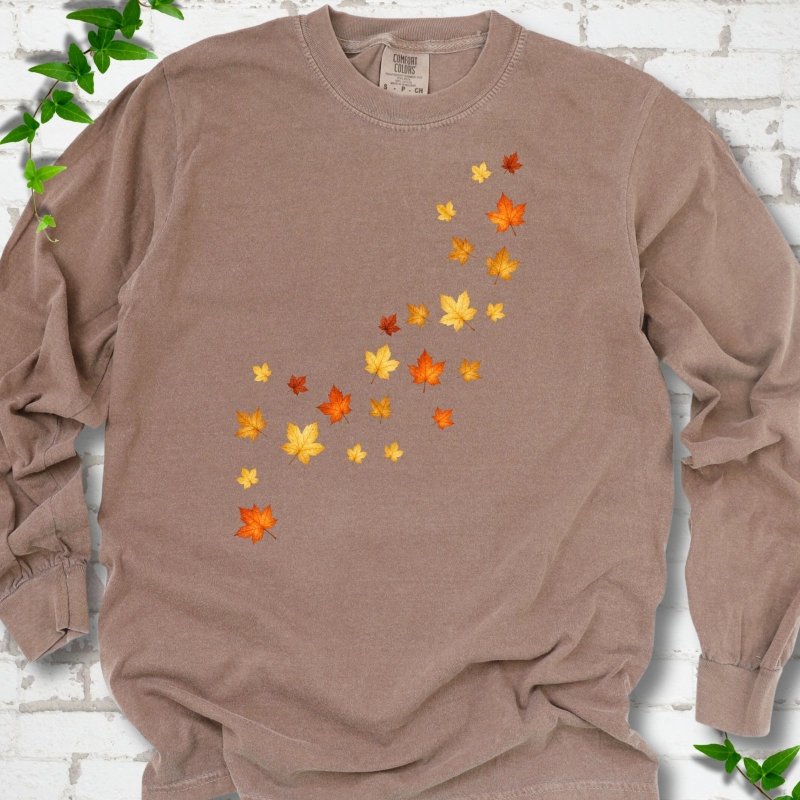 Espresso brown long sleeve t-shirt with a diagonal drift of small orange, gold and red maple leaves falling across the front, flat lay on white brick with ivy