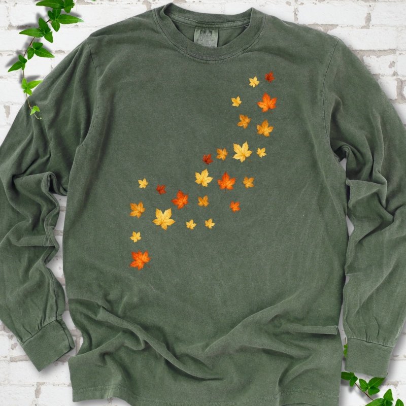 Hemp dark green long sleeve t-shirt with a diagonal drift of small orange, gold and red maple leaves falling across the front, flat lay on white brick with ivy