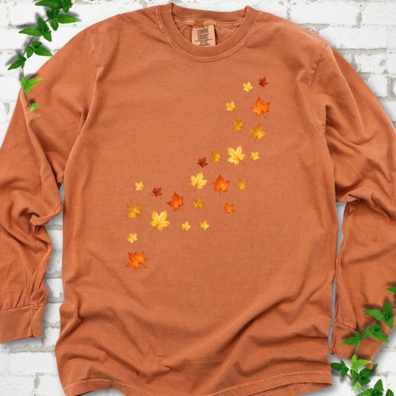 Yam burnt orange long sleeve t-shirt with a diagonal drift of small orange, gold and red maple leaves falling across the front, flat lay on white brick with ivy