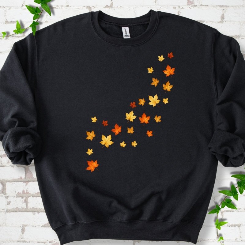 Black crewneck sweatshirt with a diagonal drift of small orange, gold and red maple leaves falling across the front, flat lay on white brick with ivy