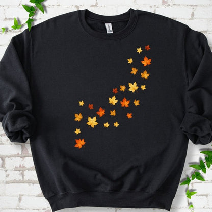 Black crewneck sweatshirt with a diagonal drift of small orange, gold and red maple leaves falling across the front, flat lay on white brick with ivy