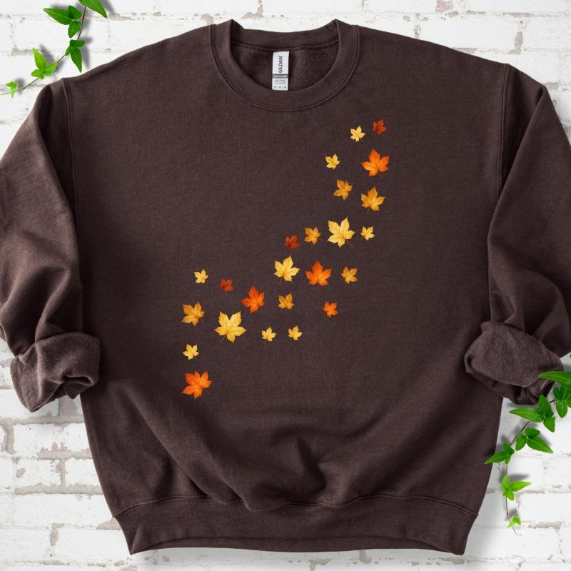 Dark Chocolate brown crewneck sweatshirt with a diagonal drift of small orange, gold and red maple leaves falling across the front, flat lay on white brick with ivy