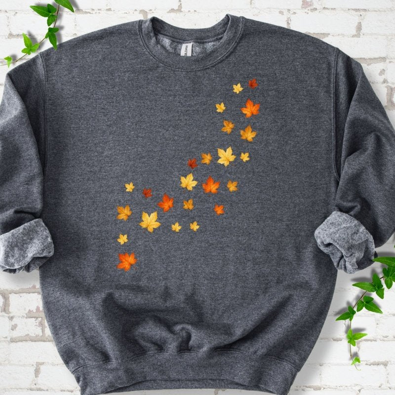 Dark Heather gray crewneck sweatshirt with a diagonal drift of small orange, gold and red maple leaves falling across the front, flat lay on white brick with ivy