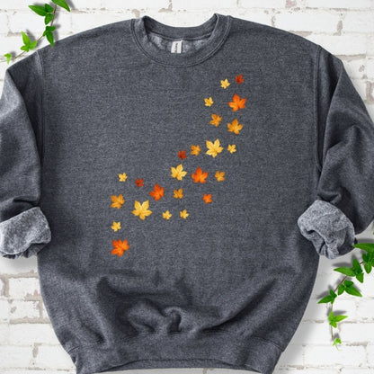 Dark Heather gray crewneck sweatshirt with a diagonal drift of small orange, gold and red maple leaves falling across the front, flat lay on white brick with ivy