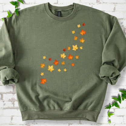 Military olive green crewneck sweatshirt with a diagonal drift of small orange, gold and red maple leaves falling across the front, flat lay on white brick with ivy