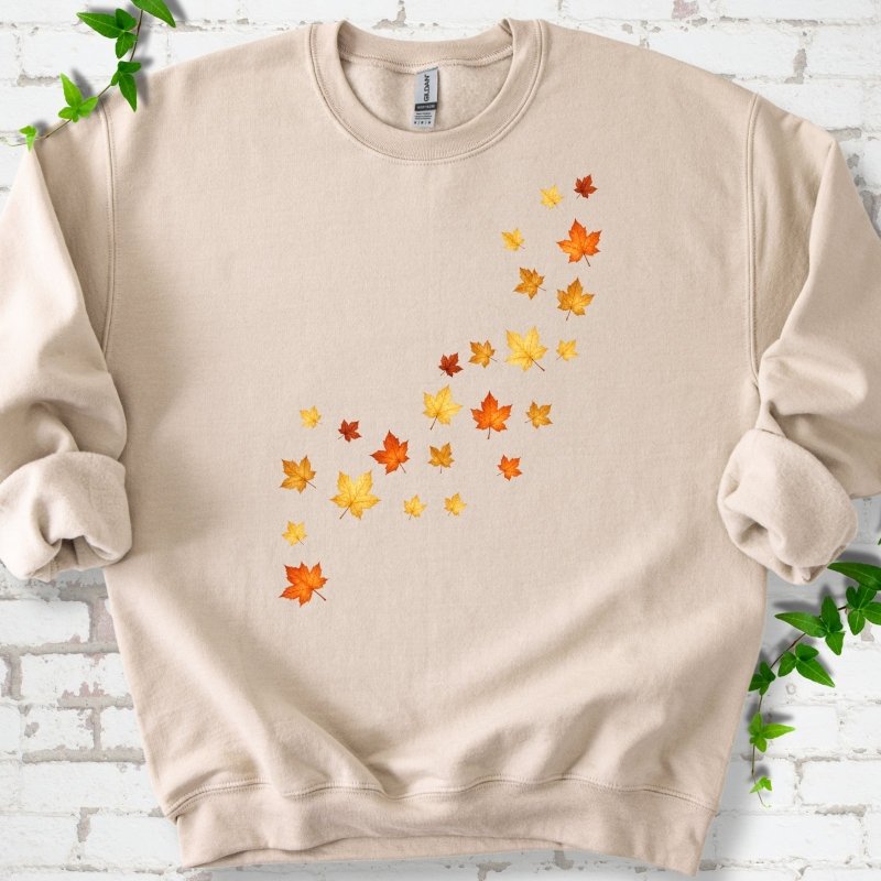 Sand beige crewneck sweatshirt with a diagonal drift of small orange, gold and red maple leaves falling across the front, flat lay on white brick with ivy