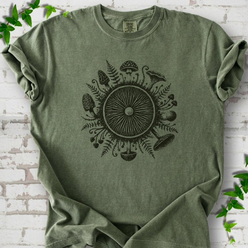 Moss green short-sleeve tee with a mushroom gill circle ringed by ferns, morels and mushrooms in dark green ink.