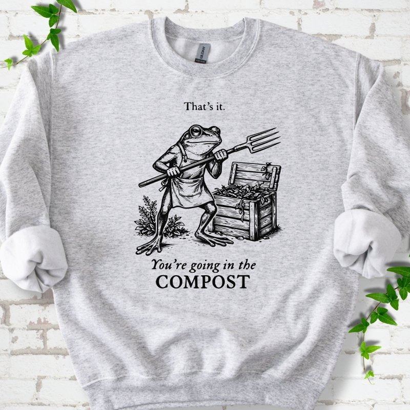 Ash classic sweatshirt laid flat, showing a frog holding a garden fork beside a compost bin and the words “That’s it. You’re going in the COMPOST.”