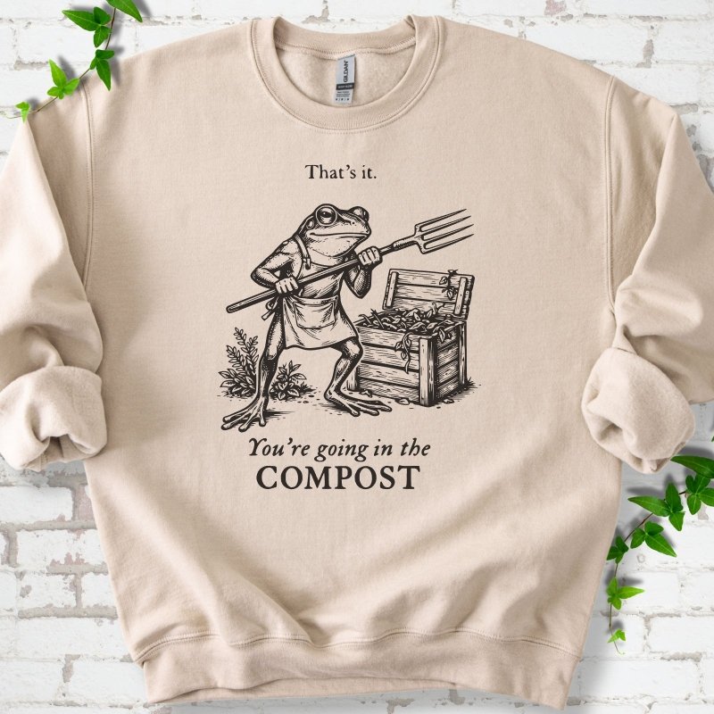 Sand classic sweatshirt laid flat, showing a frog holding a garden fork beside a compost bin and the words “That’s it. You’re going in the COMPOST.”