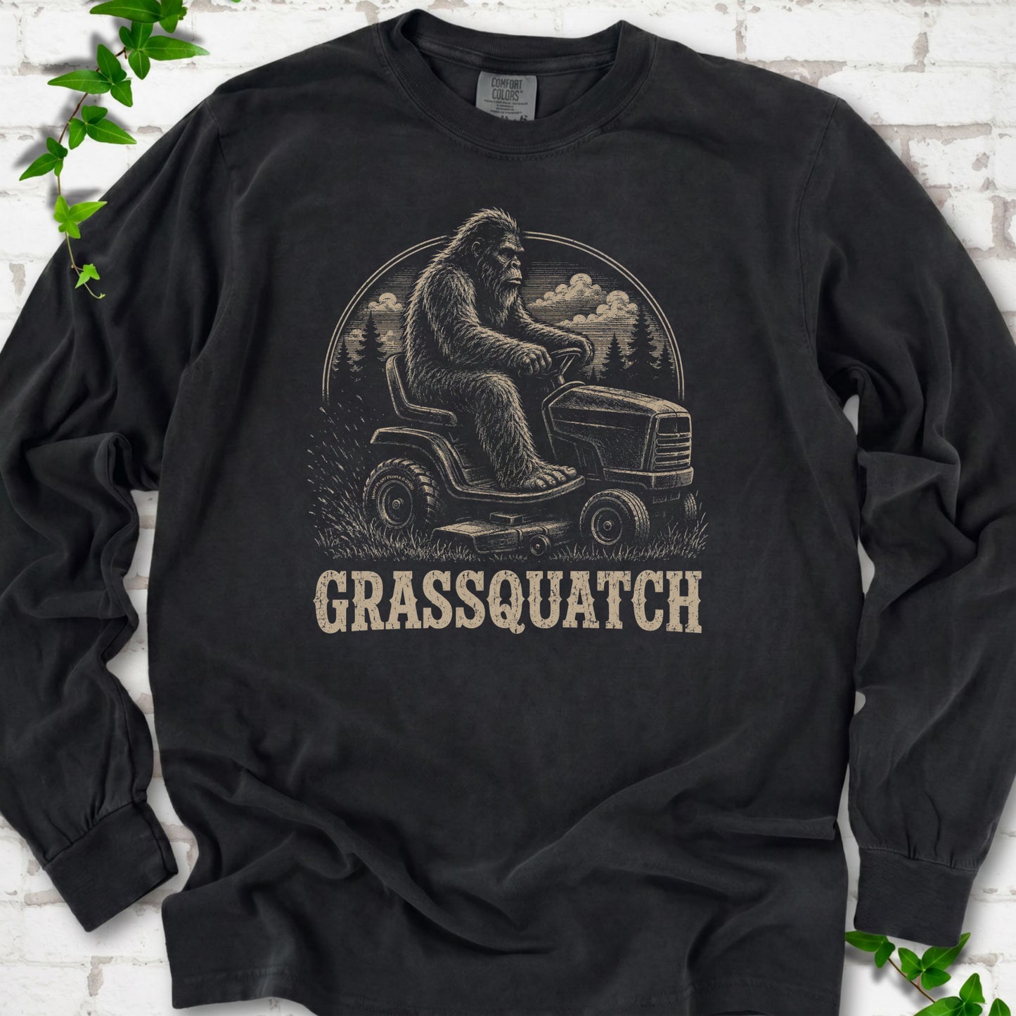 Black Comfort Colors® long sleeve T-shirt front mockup featuring Bigfoot riding a lawn mower above the word Grassquatch.