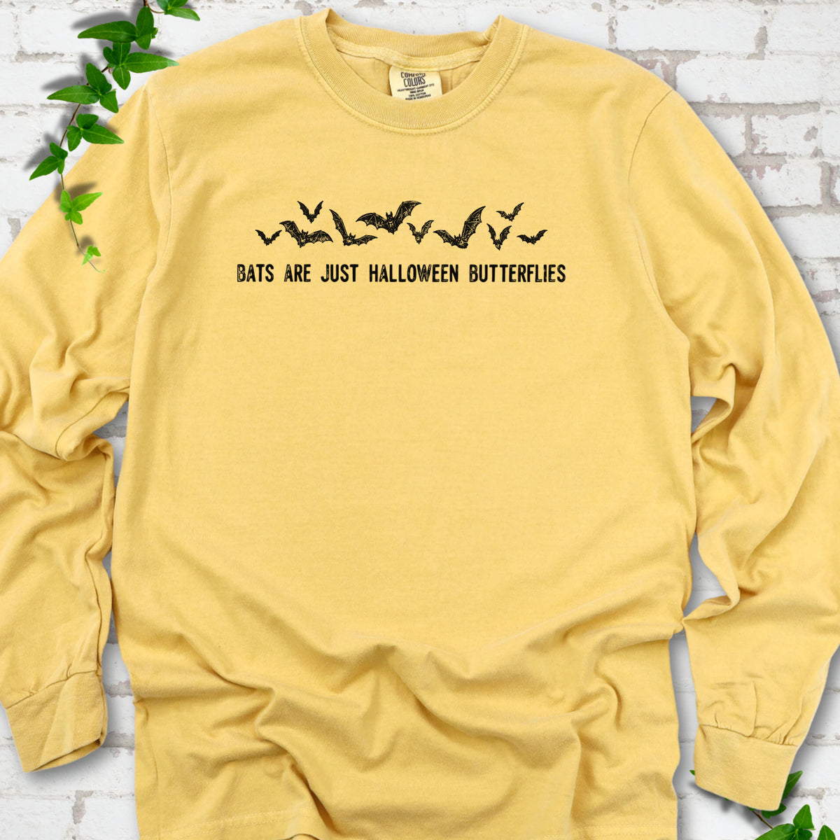 Mustard Comfort Colors® long sleeve tee flat lay with black flying bats and “Bats Are Just Halloween Butterflies” lettering.