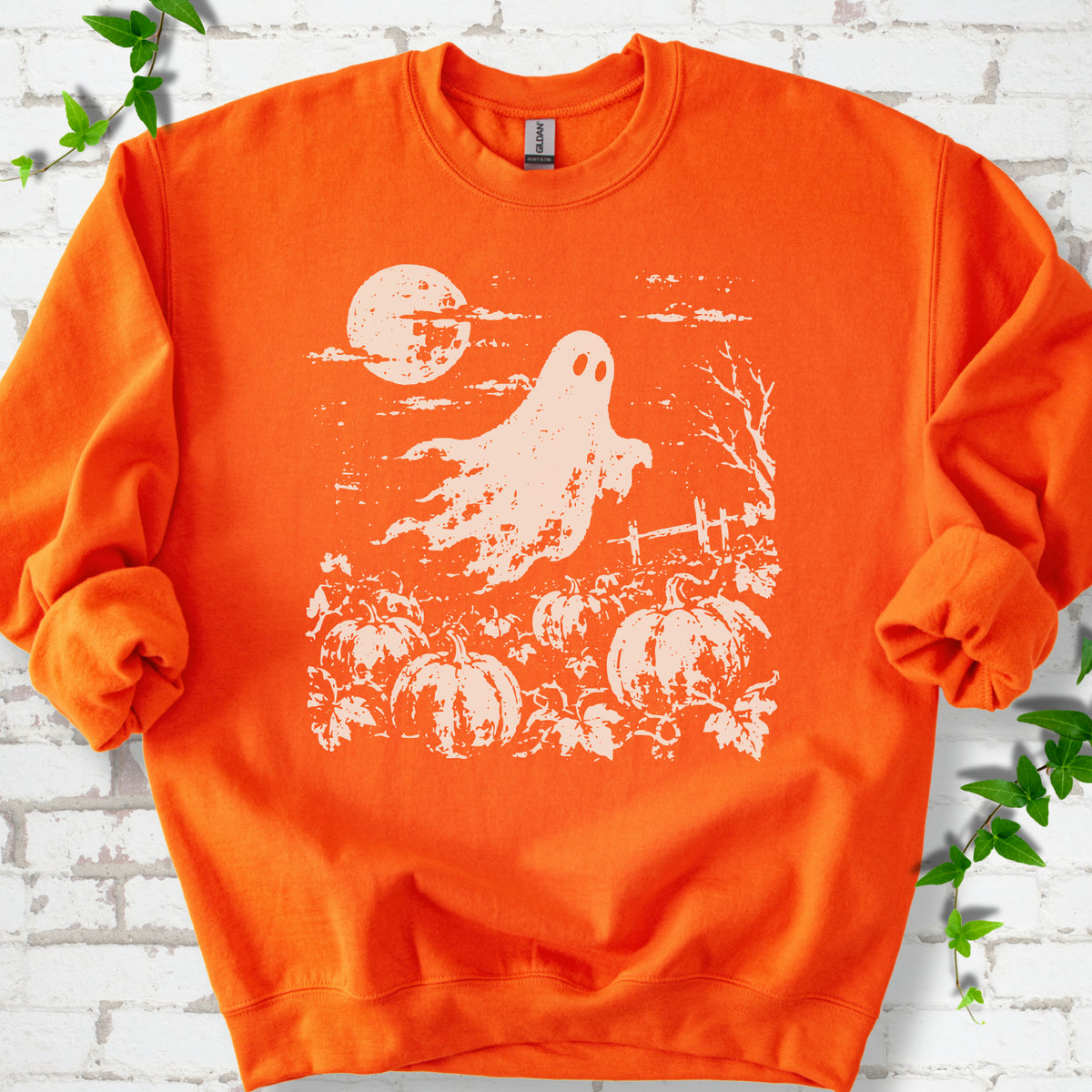 Orange crewneck sweatshirt flat lay featuring a distressed ghost floating above a pumpkin patch beneath a full moon.