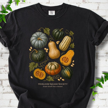 Black Comfort Colors® short sleeve T-shirt flat lay featuring an illustrated collection of heirloom squash, pumpkins, seeds and vines with text "Heirloom Squash Society" and "Every shape has a season"