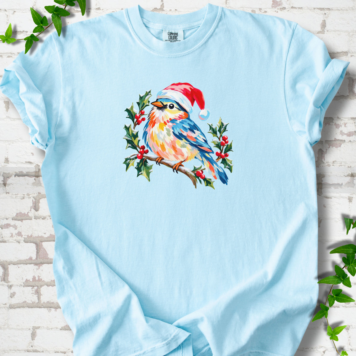 Chambray T-shirt laid flat, featuring a colorful songbird in a red festive hat perched among holly leaves and berries.