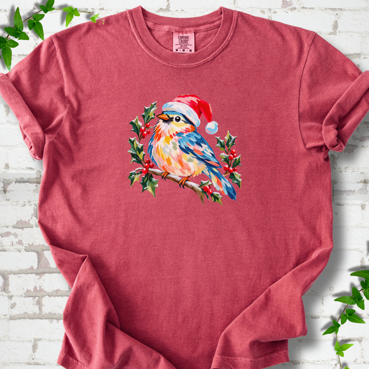 Crimson T-shirt laid flat, featuring a colorful songbird in a red festive hat perched among holly leaves and berries.