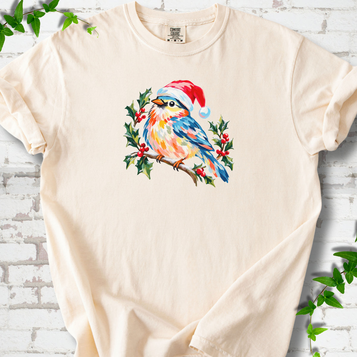 Ivory T-shirt laid flat, featuring a colorful songbird in a red festive hat perched among holly leaves and berries.