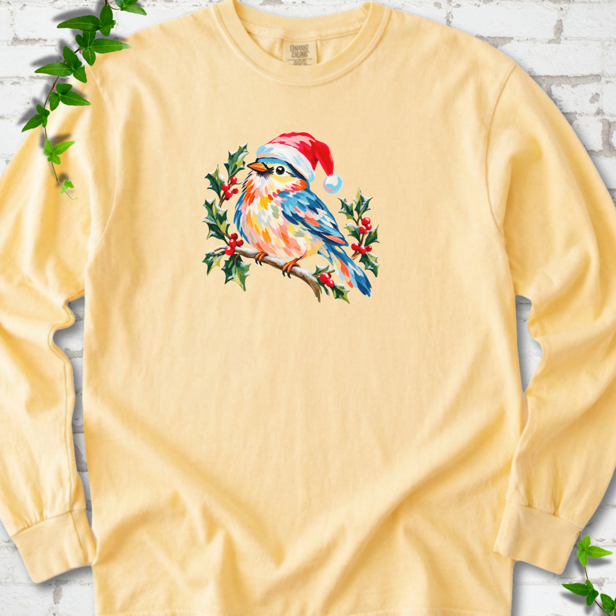 Butter long sleeve T-shirt laid flat, featuring a colorful songbird in a red festive hat perched among holly leaves and berries.