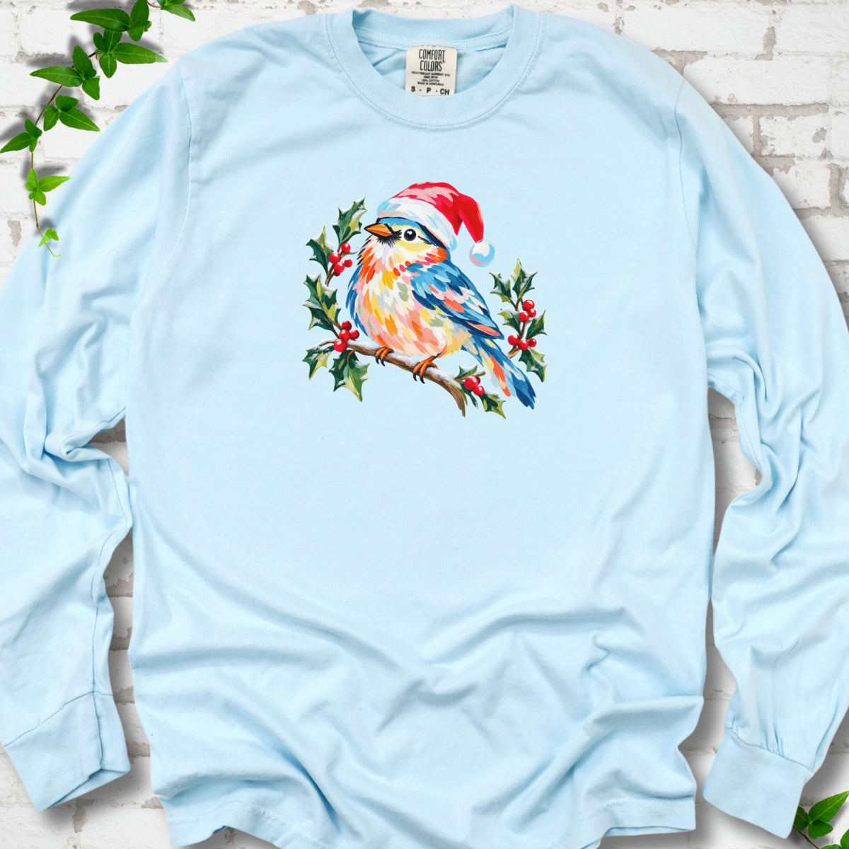 Chambray long sleeve T-shirt laid flat, featuring a colorful songbird in a red festive hat perched among holly leaves and berries.
