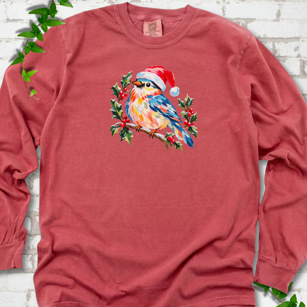 Crimson long sleeve T-shirt laid flat, featuring a colorful songbird in a red festive hat perched among holly leaves and berries.