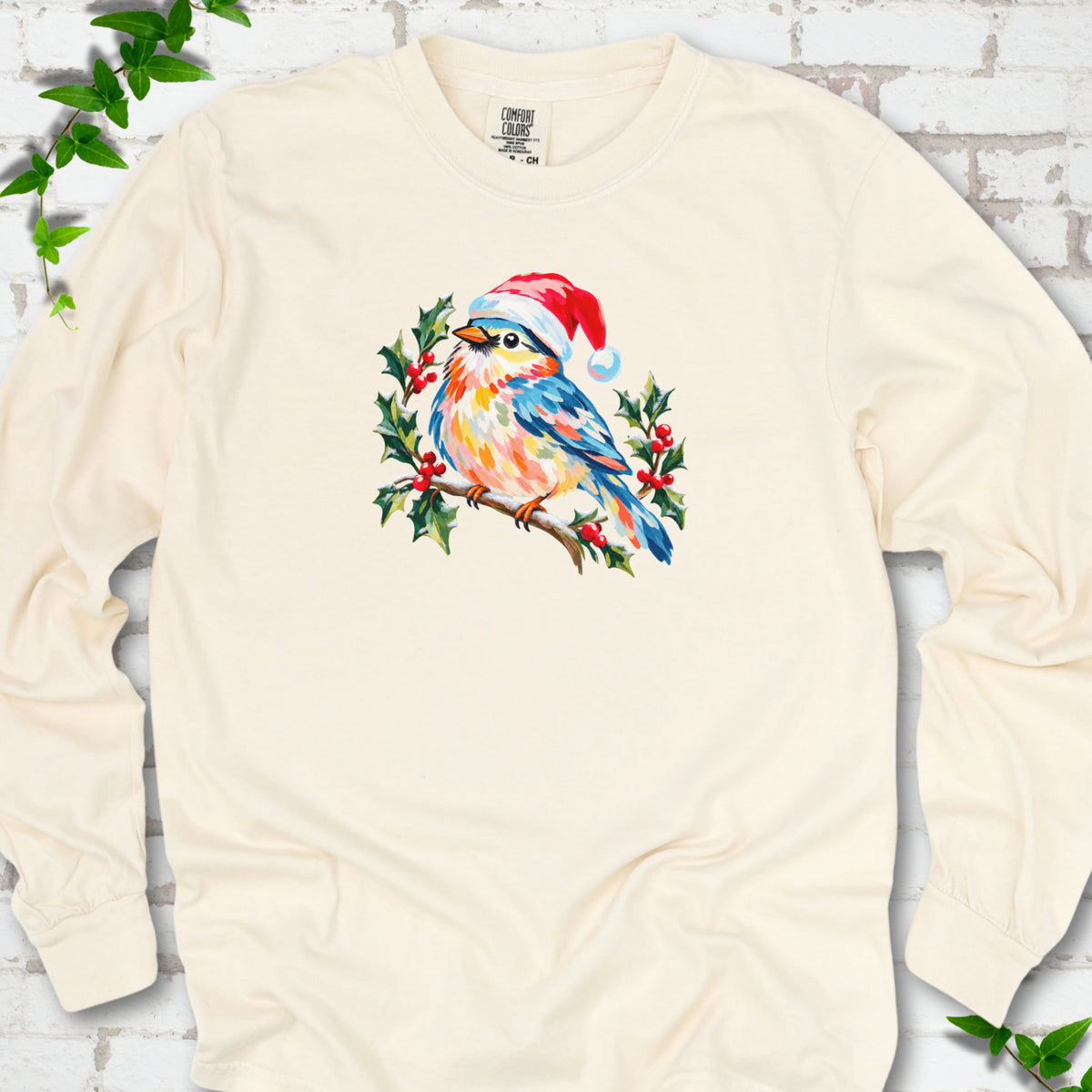 Ivory long sleeve T-shirt laid flat, featuring a colorful songbird in a red festive hat perched among holly leaves and berries.