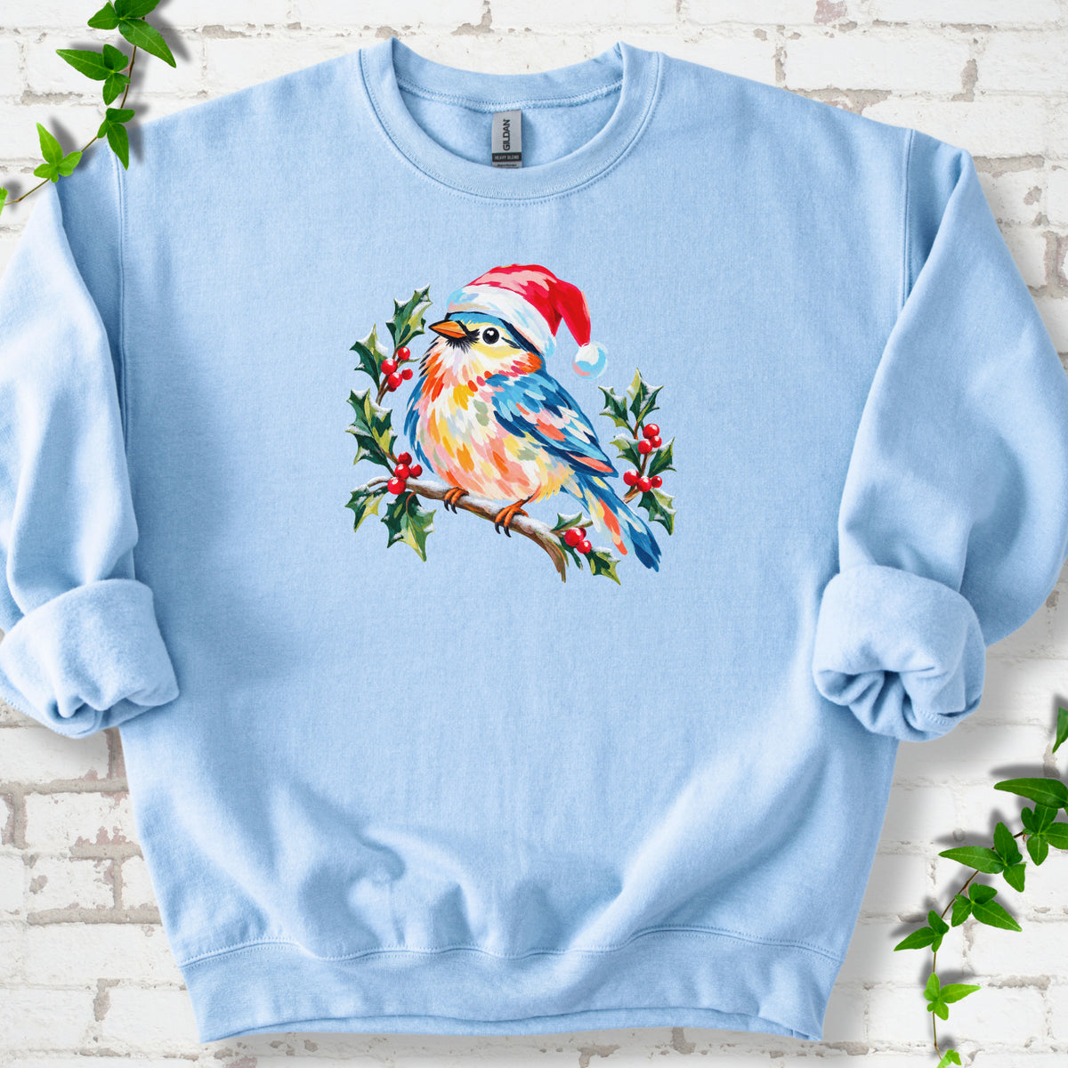 Light Blue classic sweatshirt laid flat, featuring a colorful songbird in a red festive hat perched among holly leaves and berries.