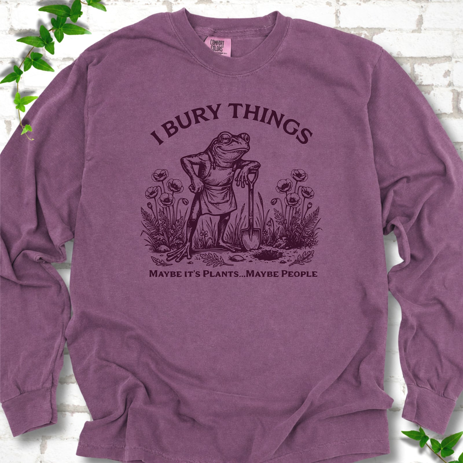 Berry Comfort Colors® long sleeve T-shirt flat lay featuring a frog with a shovel in a flower garden and the words I Bury Things.