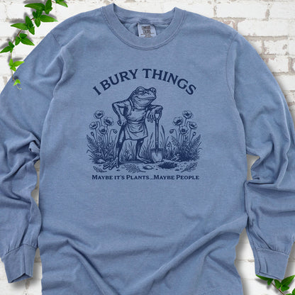 Blue Jean Comfort Colors® long sleeve T-shirt flat lay featuring a frog with a shovel in a flower garden and the words I Bury Things.