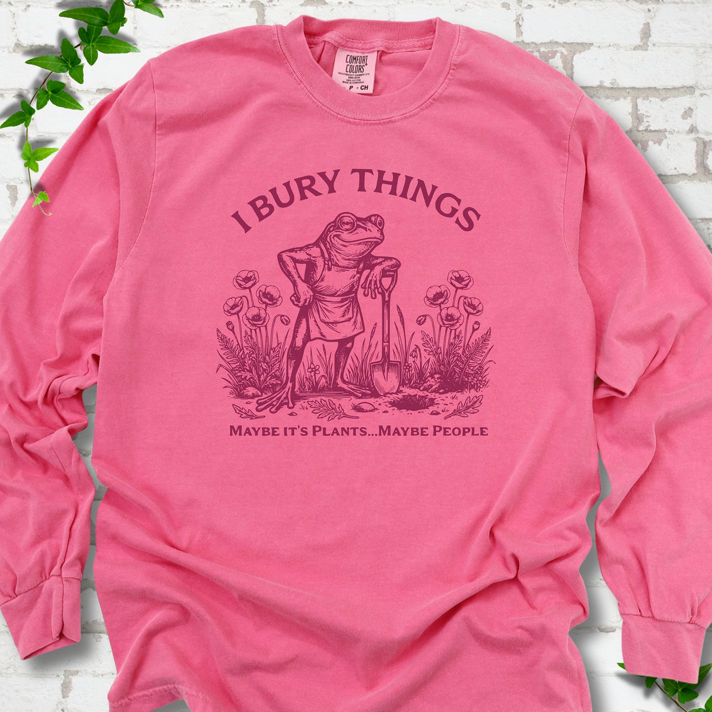 Crunchberry Comfort Colors® long sleeve T-shirt flat lay featuring a frog with a shovel in a flower garden and the words I Bury Things.