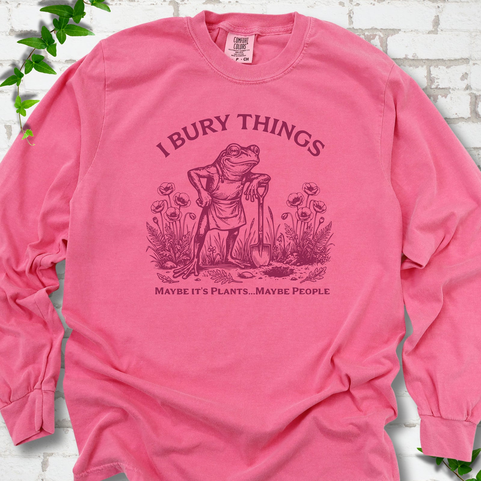 Crunchberry Comfort Colors® long sleeve T-shirt flat lay featuring a frog with a shovel in a flower garden and the words I Bury Things.