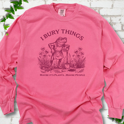 Crunchberry Comfort Colors® long sleeve T-shirt flat lay featuring a frog with a shovel in a flower garden and the words I Bury Things.