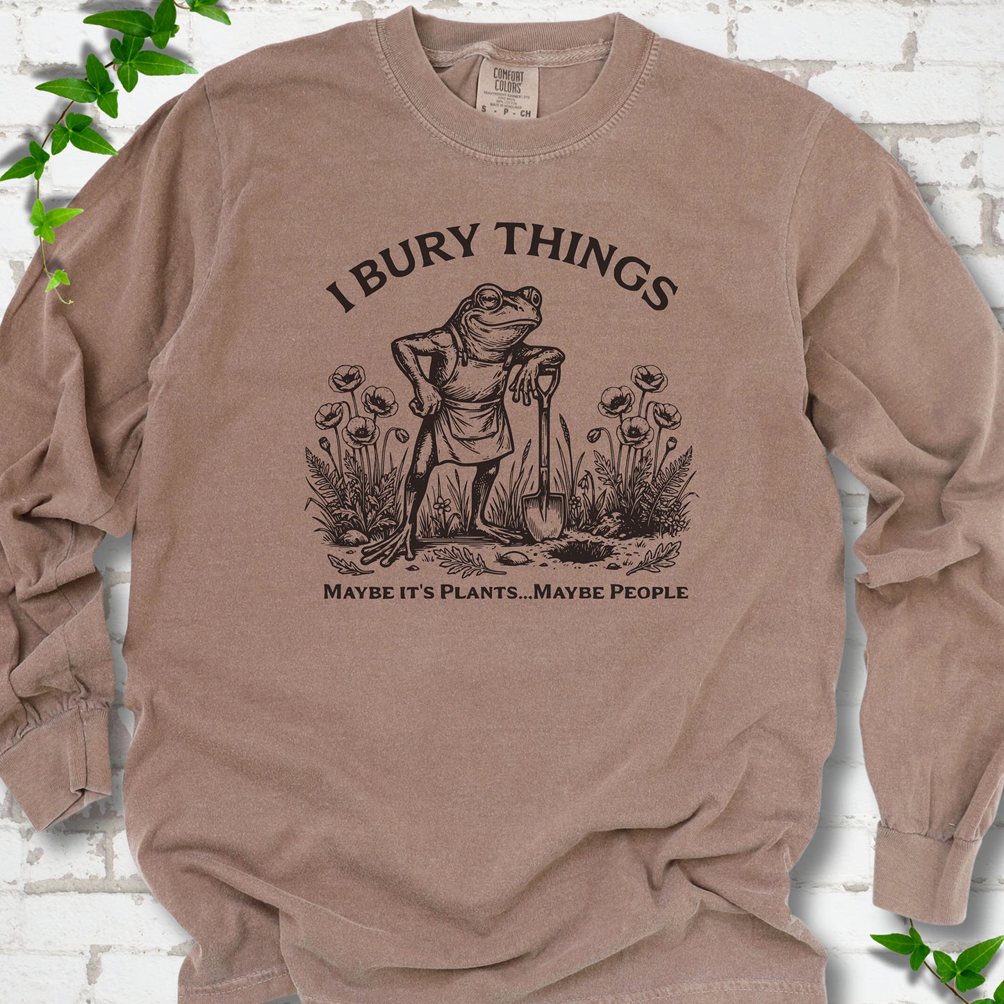 Espresso Comfort Colors® long sleeve T-shirt flat lay featuring a frog with a shovel in a flower garden and the words I Bury Things.