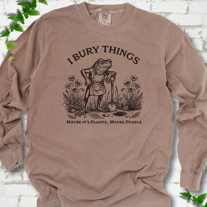 Espresso Comfort Colors® long sleeve T-shirt flat lay featuring a frog with a shovel in a flower garden and the words I Bury Things.