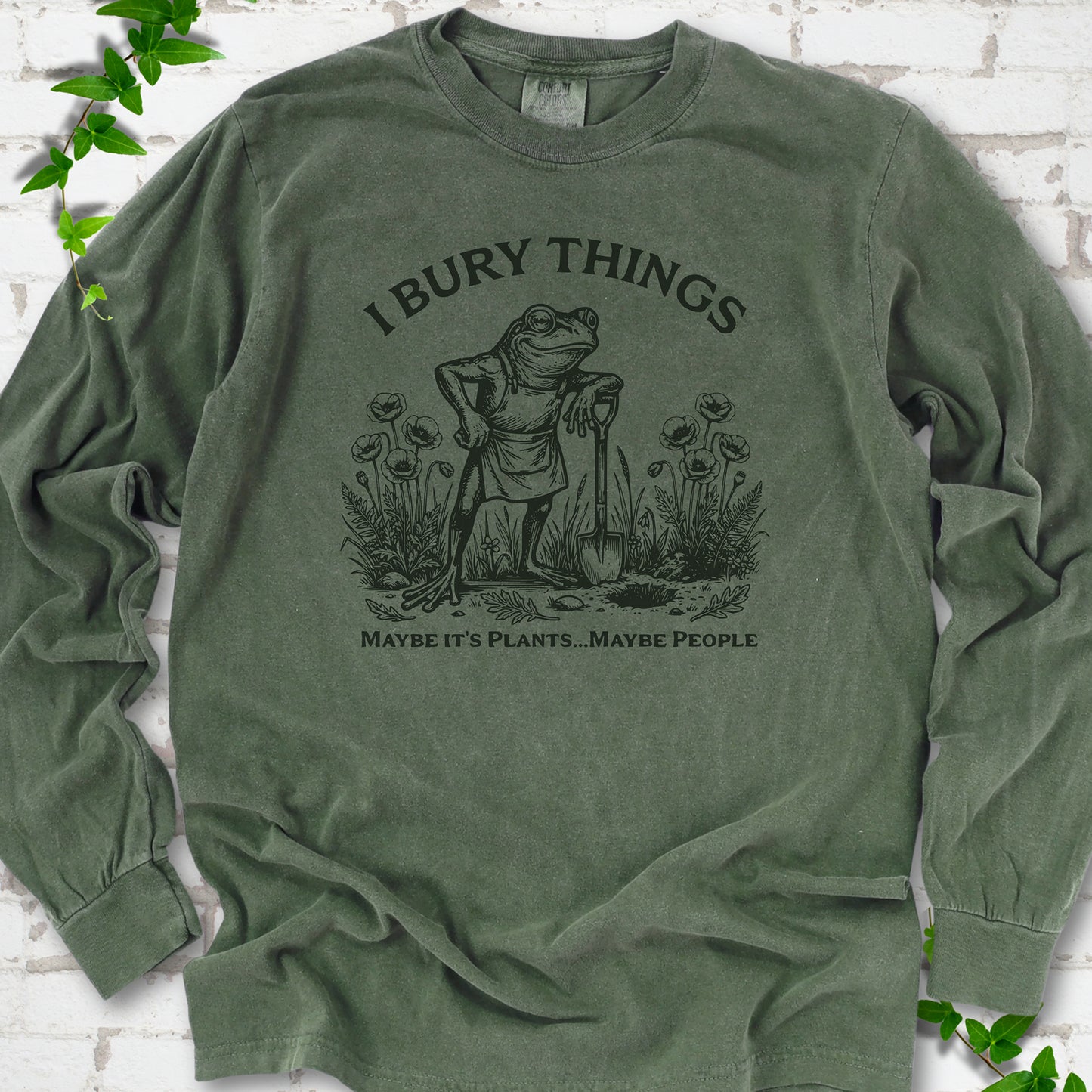 Hemp Comfort Colors® long sleeve T-shirt flat lay featuring a frog with a shovel in a flower garden and the words I Bury Things.