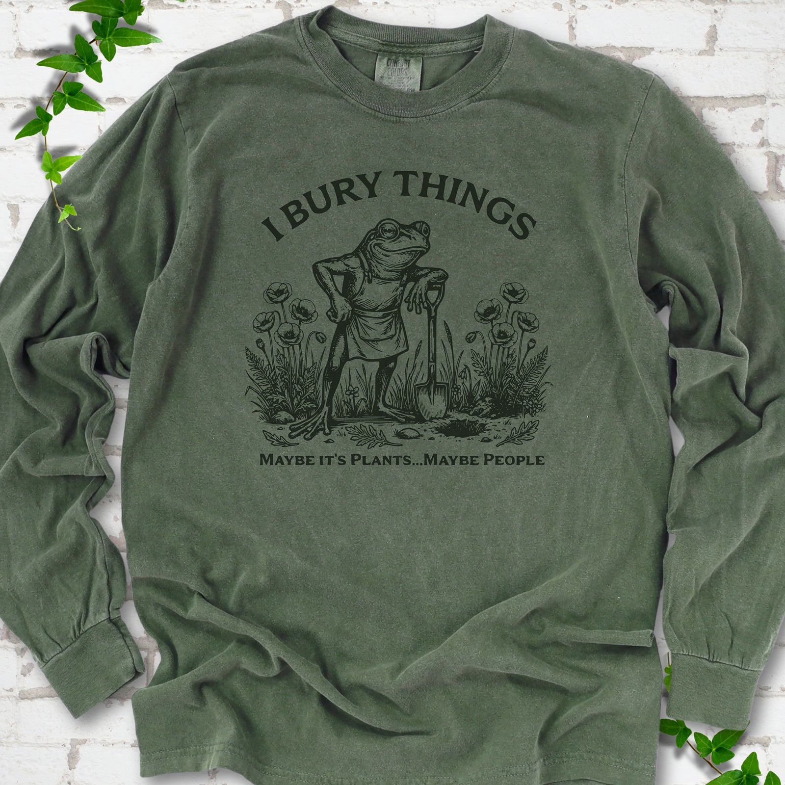 Hemp Comfort Colors® long sleeve T-shirt flat lay featuring a frog with a shovel in a flower garden and the words I Bury Things.