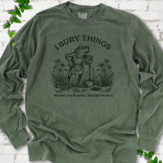 Hemp Comfort Colors® long sleeve T-shirt flat lay featuring a frog with a shovel in a flower garden and the words I Bury Things.