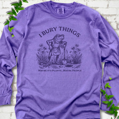 Violet Comfort Colors® long sleeve T-shirt flat lay featuring a frog with a shovel in a flower garden and the words I Bury Things.