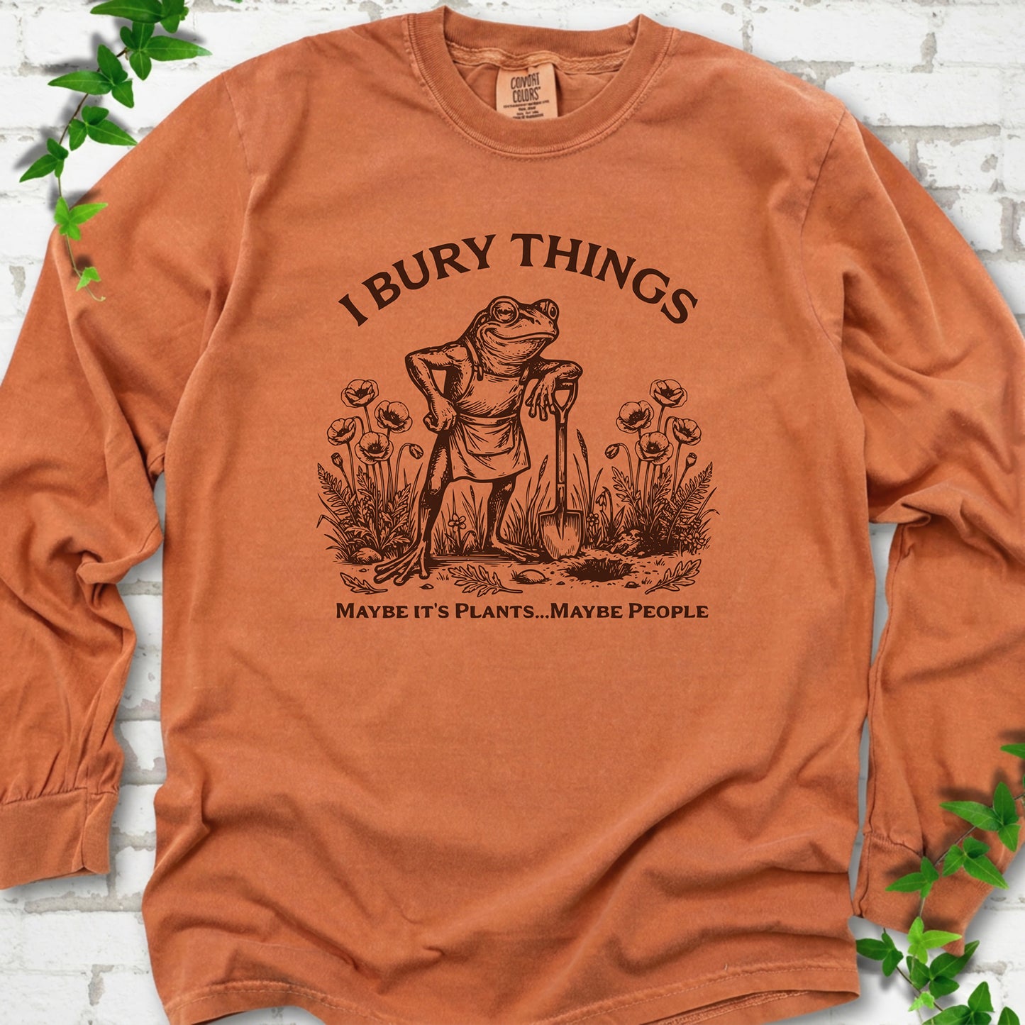 Yam Comfort Colors® long sleeve T-shirt flat lay featuring a frog with a shovel in a flower garden and the words I Bury Things.