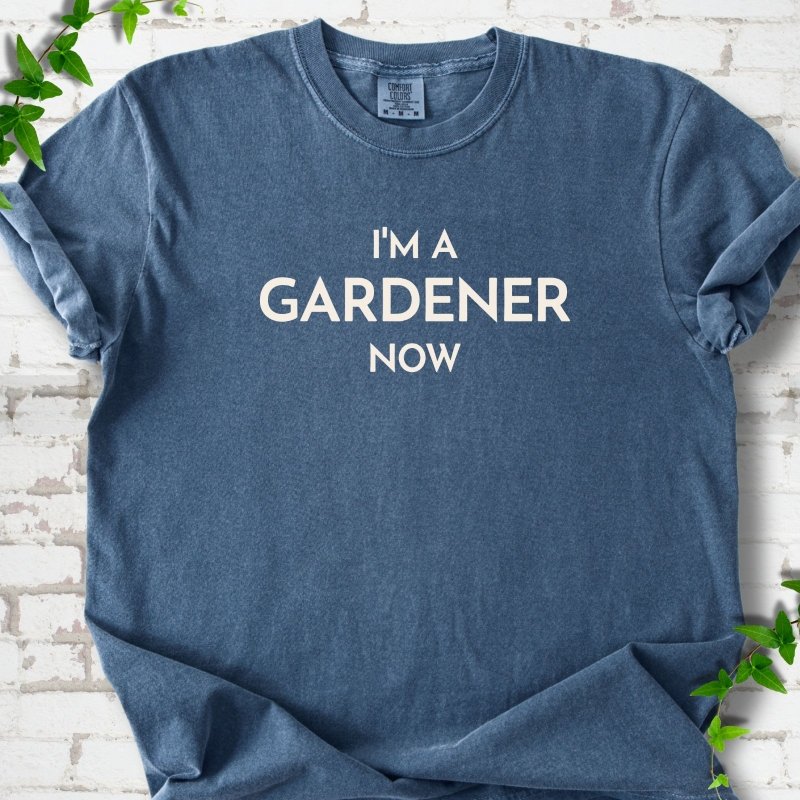 Denim short sleeve T-shirt laid flat on white brick with greenery, showing the cream words 'I'm a Gardener Now' centered on the chest.