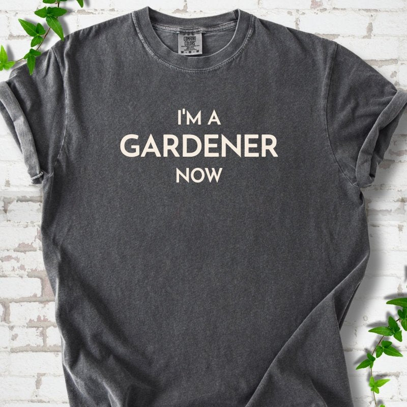 Pepper short sleeve T-shirt laid flat on white brick with greenery, showing the cream words 'I'm a Gardener Now' centered on the chest.