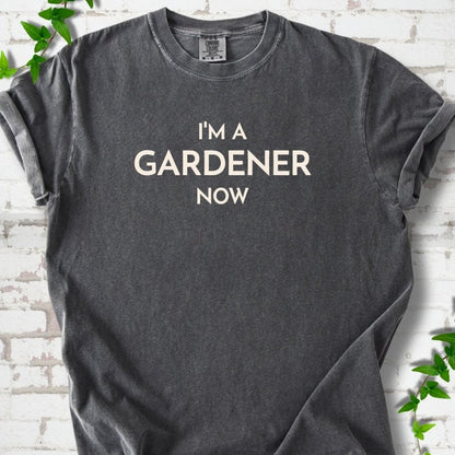 Pepper short sleeve T-shirt laid flat on white brick with greenery, showing the cream words 'I'm a Gardener Now' centered on the chest.